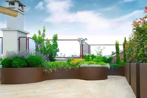 Composizioni fioriere da esterno con piante verdi e fiori colorati su una terrazza pavimentata, sotto un cielo azzurro con nuvole bianche.